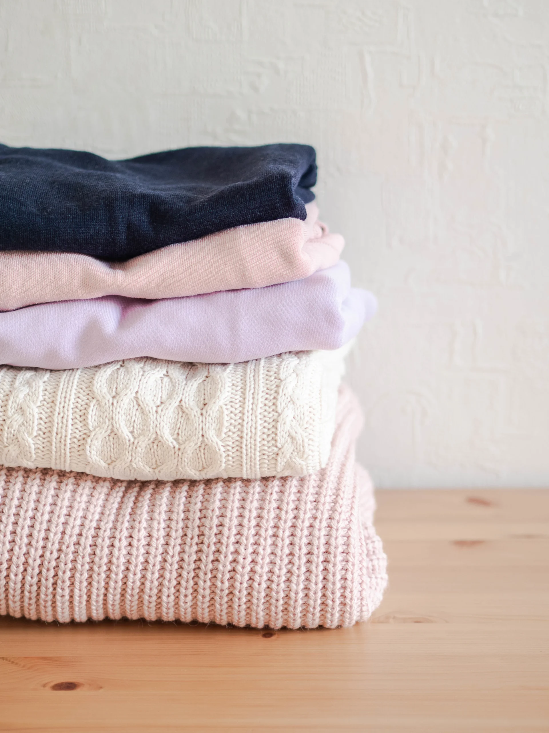 Stack of warm knitted woolen sweaters pastel shades and dark blue on a wood table